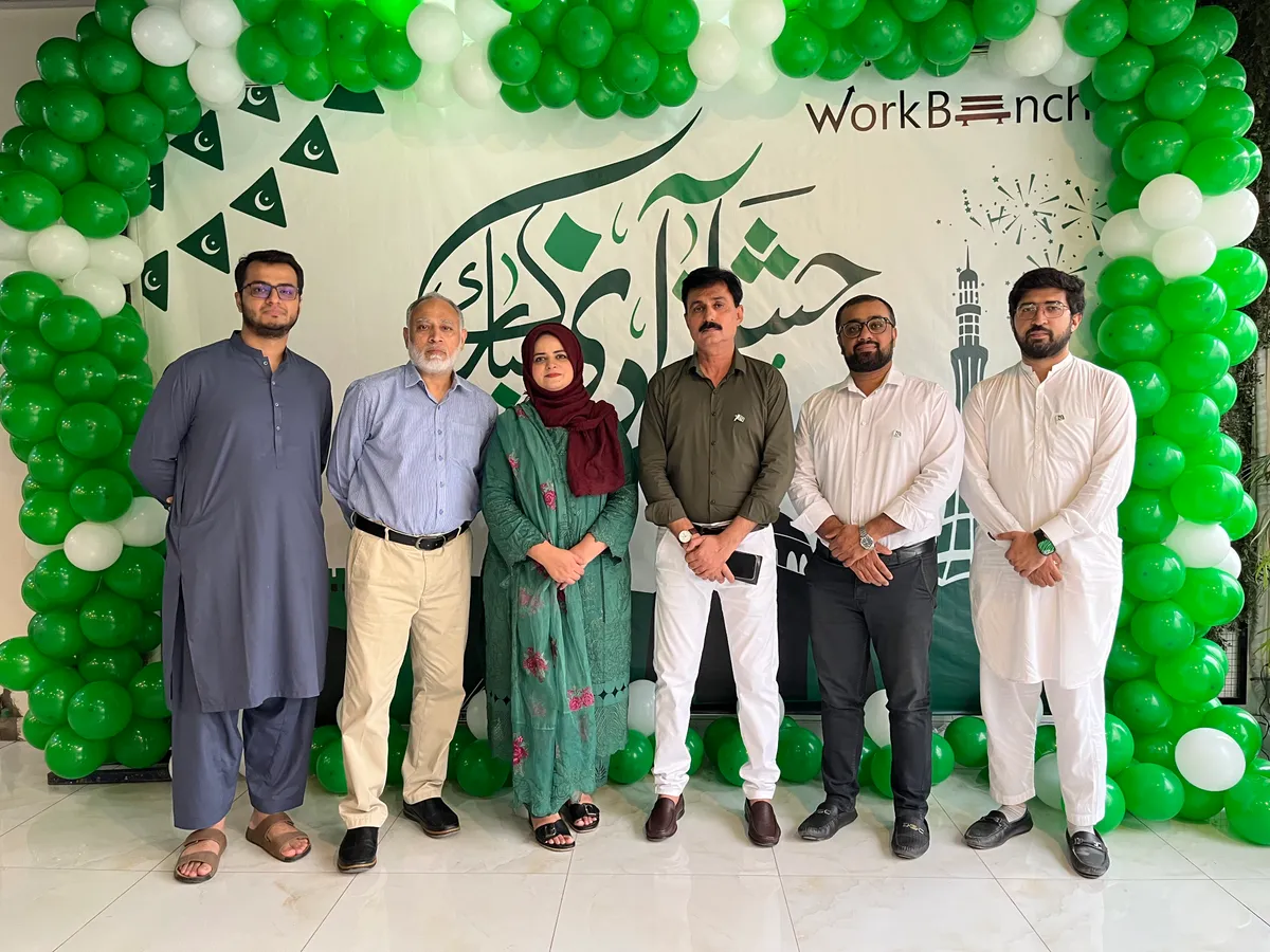 Members standing together in front of a green and white Independence Day backdrop at WorkBench, with Jashn e Azadi calligraphy, a Minar e Pakistan illustration and an arch of green and white balloons.