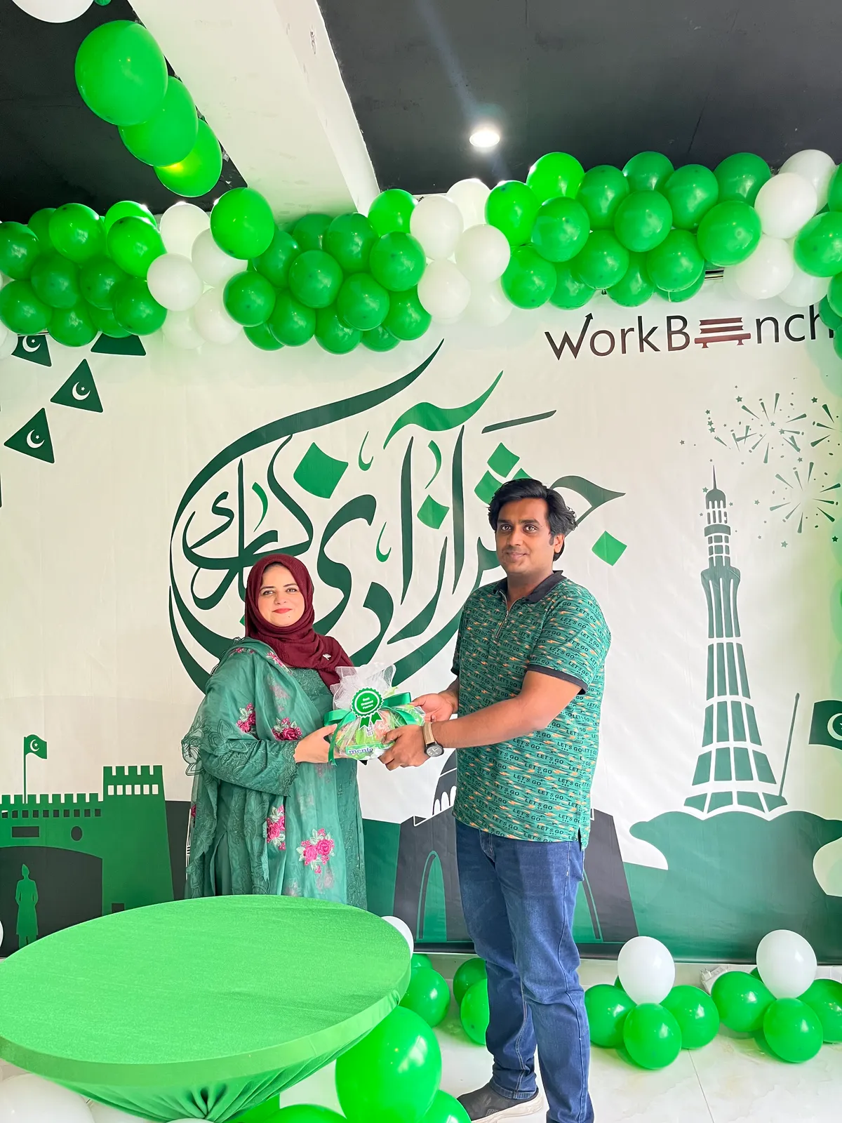 A member handing a green and white wrapped gift to a colleague in front of the Independence Day backdrop at WorkBench, under an arch of green and white balloons.