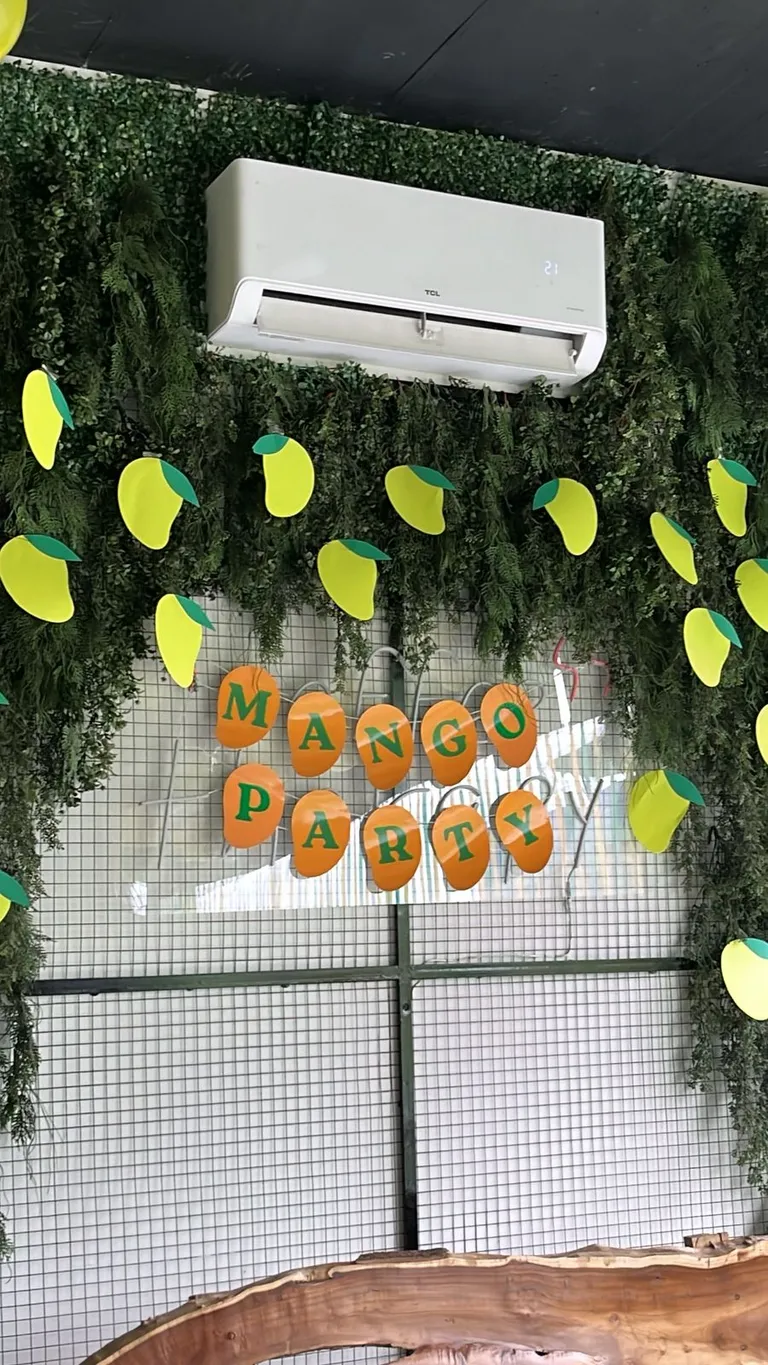 Mango Party banner of orange paper mangoes on a green foliage wall