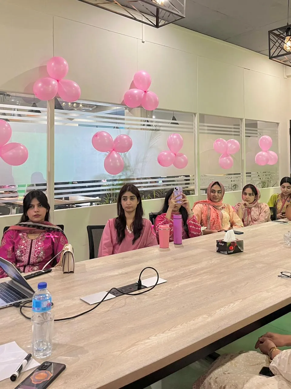 Women seated along one side of the long meeting room table at WorkBench listening to a talk, with clusters of pink balloons taped to the glass partition behind them.