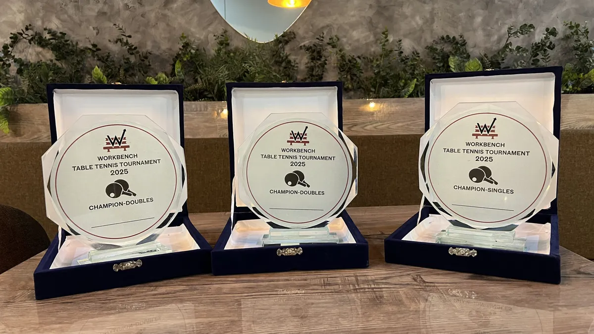 Three glass plaques in navy presentation boxes on a wooden table at WorkBench, each engraved WorkBench Table Tennis Tournament 2025 with champion doubles and champion singles titles.