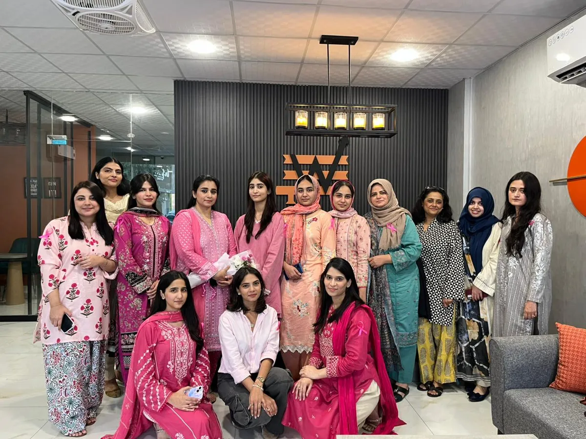 A group of women in pink and brightly coloured shalwar kameez posing together for a group photograph in the WorkBench lobby, under the amber W on the charcoal fluted wall.