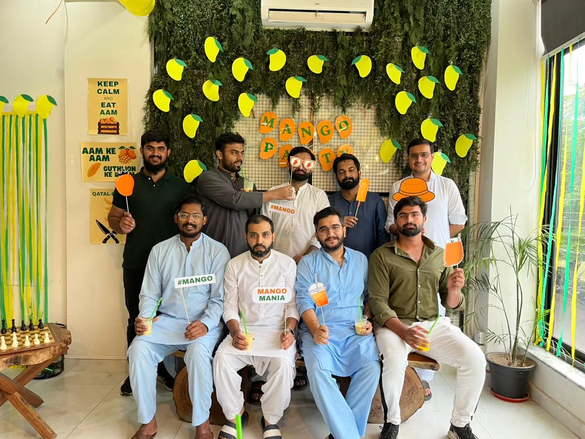 A group of members posing together at the WorkBench mango party, holding cardboard mango and mango drink props, in front of a green plant wall hung with paper mangoes and a Mango Party sign.
