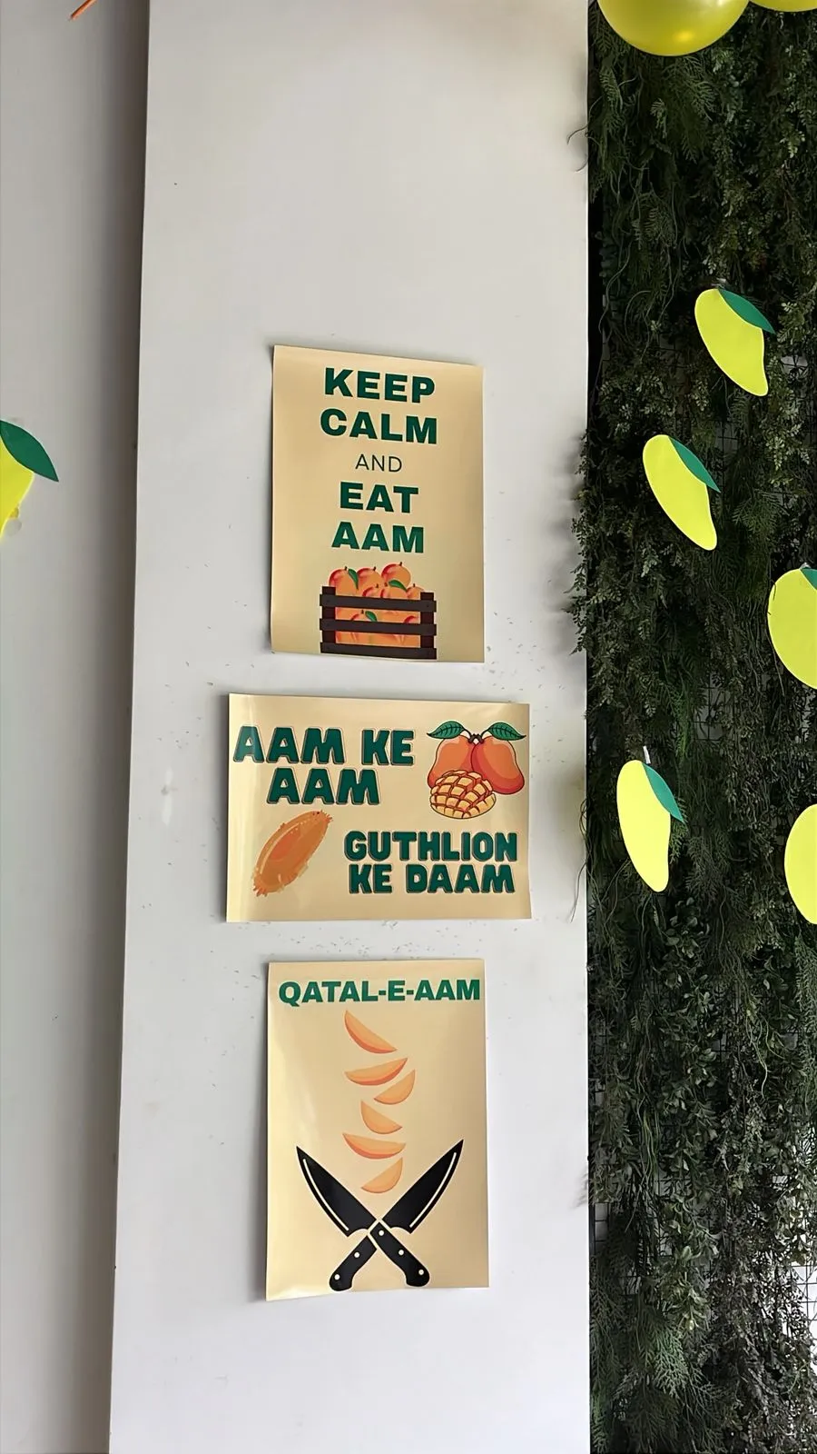 Three cream posters taped to a white wall at the WorkBench mango party reading Keep Calm and Eat Aam, Aam ke Aam Guthlion ke Daam, and Qatal e Aam, beside a plant wall hung with paper mangoes.