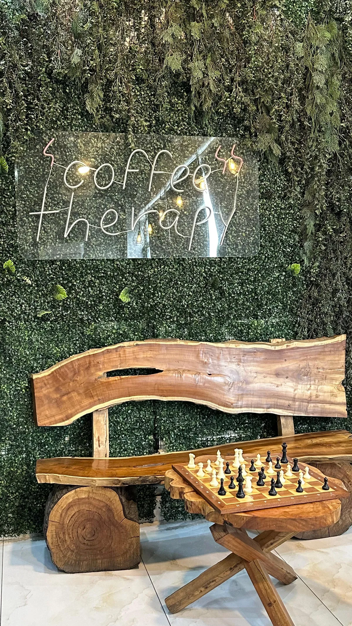 The coffee corner at WorkBench, with a dense green plant wall, a coffee therapy sign in wire script, a live edge wooden bench and a chess set on a carved wooden table.
