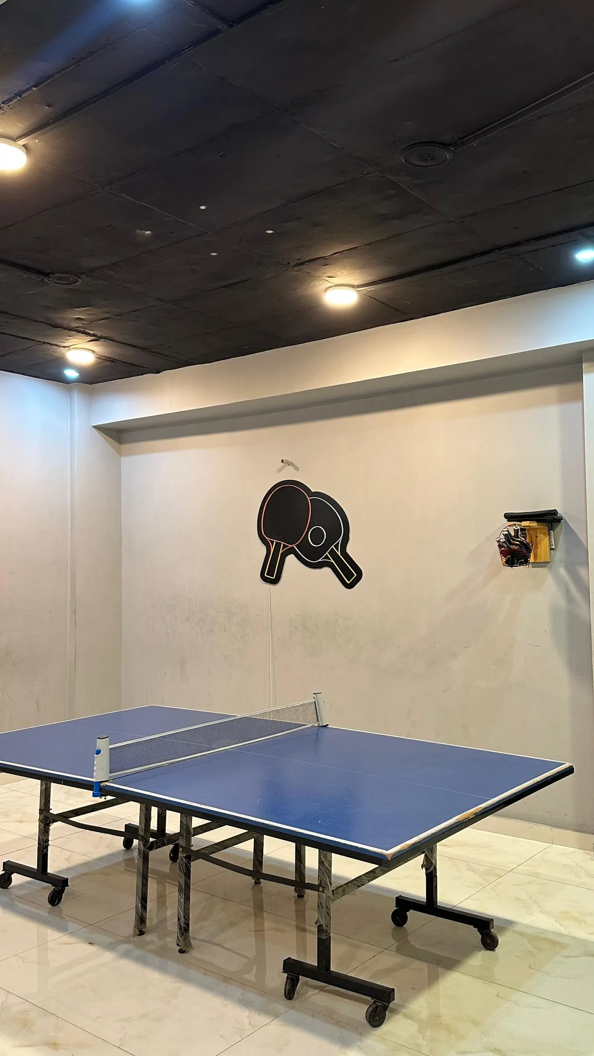 The game room at WorkBench: a blue table tennis table on a tiled floor under a black ceiling, with a painted paddle logo and a bat rack on the wall.