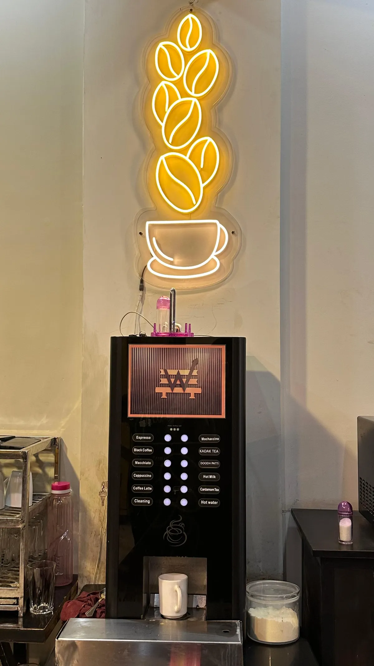 A neon sign of coffee beans falling into a cup, lit amber and white above a coffee machine at WorkBench with buttons for espresso, cappuccino, latte, kadak tea and doodh patti.