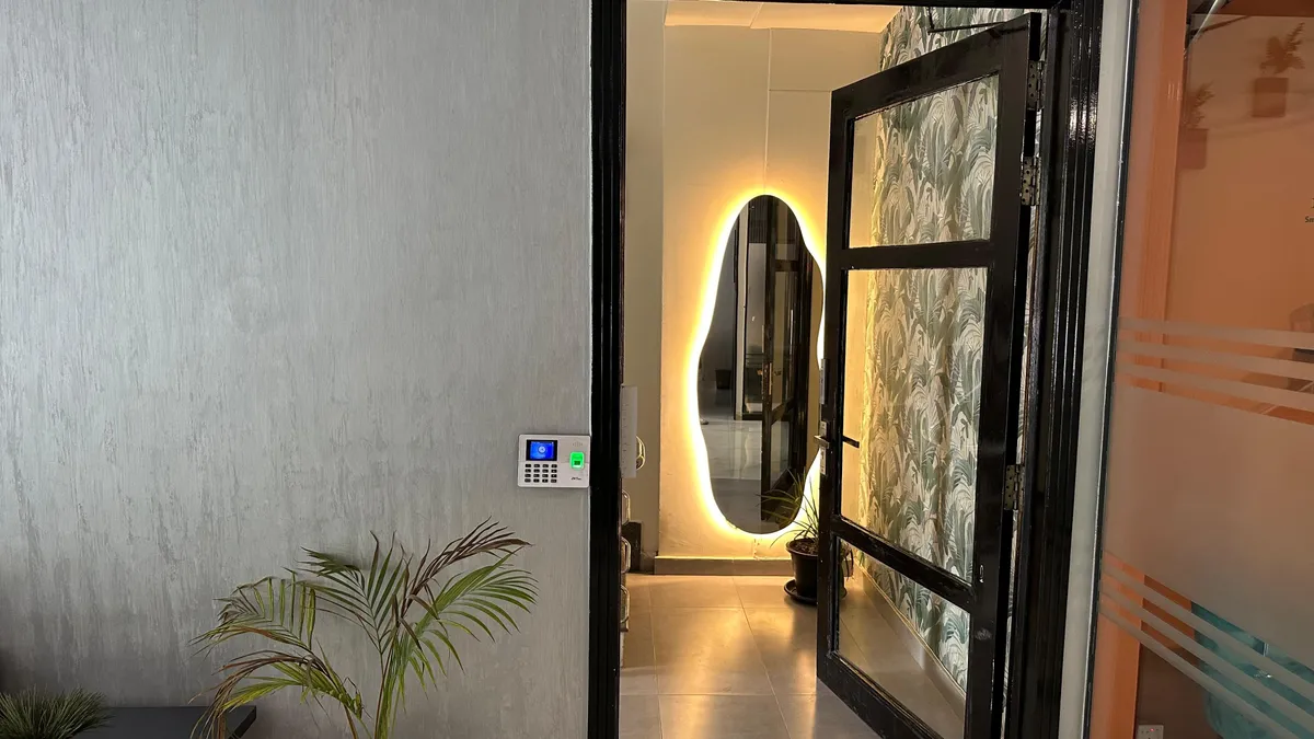 A doorway inside WorkBench opening onto a warmly backlit oval mirror, with an access keypad beside the frame and a palm in the corner.