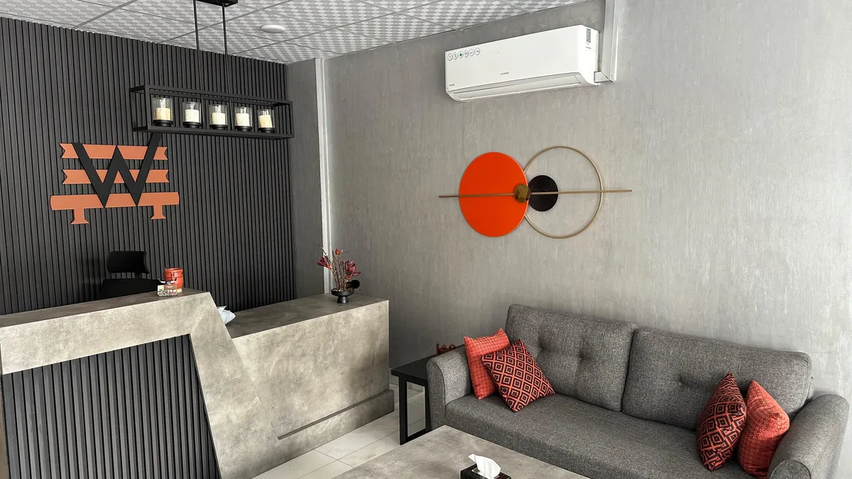 Reception at the WorkBench coworking space in Valencia Town, Lahore: a poured concrete desk beside a charcoal fluted wall carrying the amber W mark, with a grey sofa and terracotta cushions in the waiting corner.