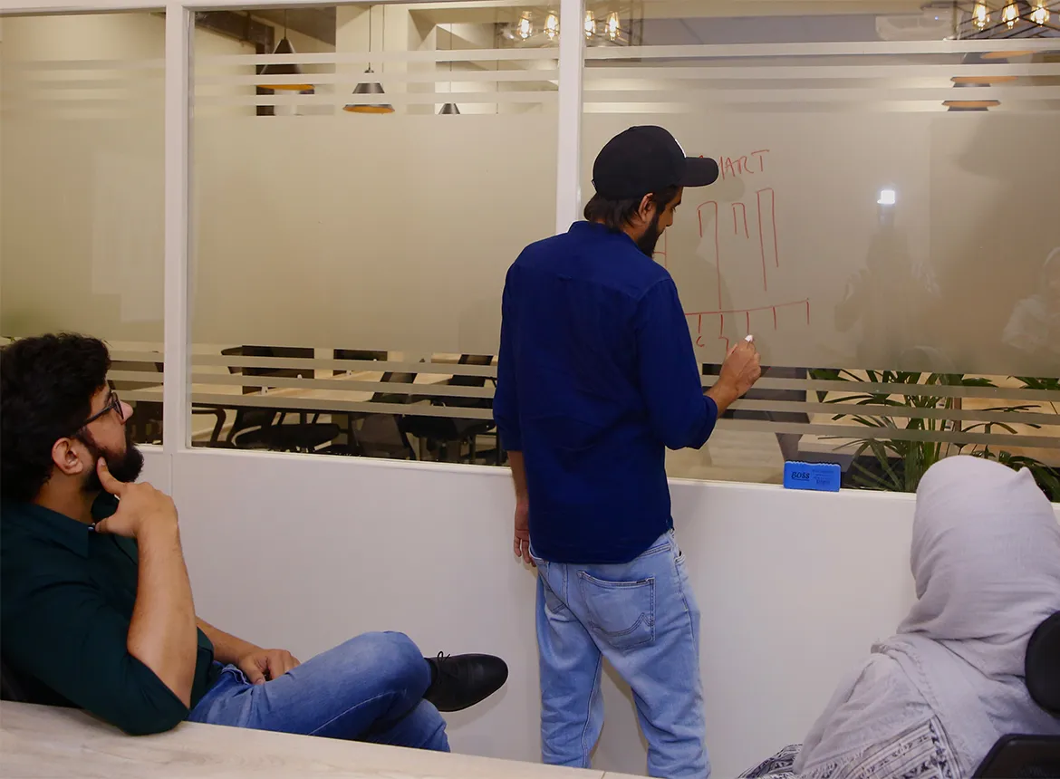 A member in a blue shirt drawing a diagram in red marker on a frosted glass wall at WorkBench while two colleagues watch from their seats.